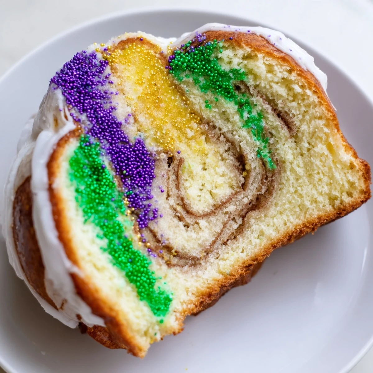 Sliced Mardi Gras King Cake showing a soft brioche texture and sweet cinnamon filling, drizzled with white icing and colorful festive sprinkles.