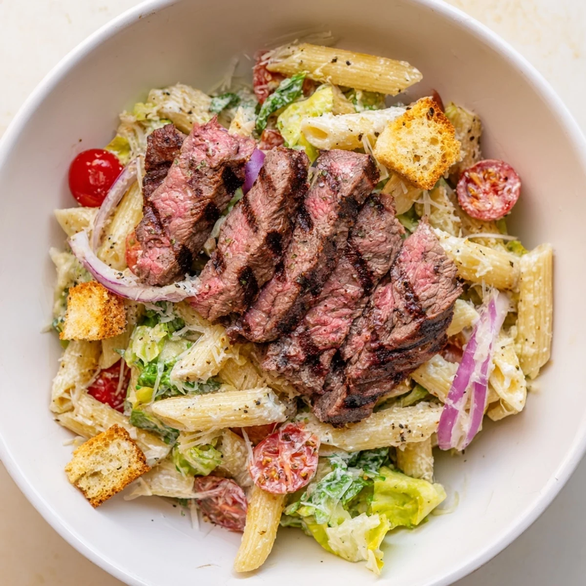 Freshly grilled steak slices top a colorful bowl of flavorful Steak Caesar Pasta Salad with crisp romaine and cherry tomatoes.