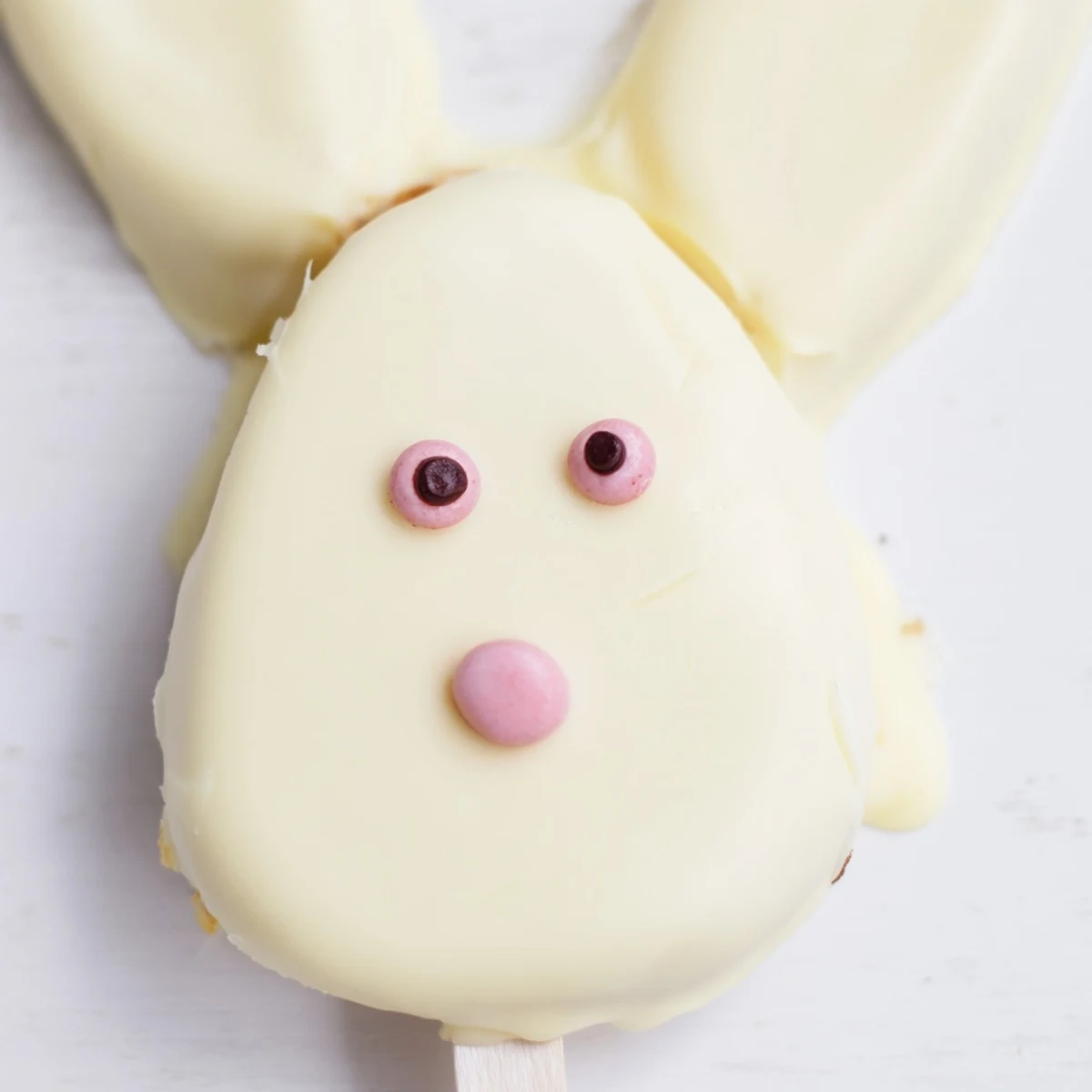 Creamy white chocolate bunny cakesicles with pink fondant ears and sprinkles displayed on parchment paper.